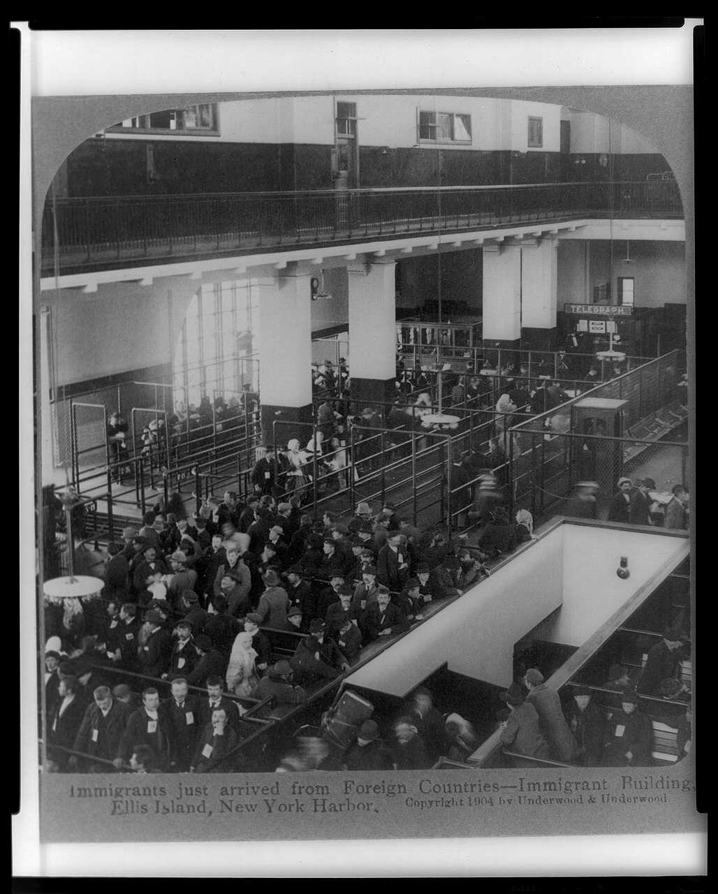 Black-and-white photograph of many people sitting on benches inside a great hall, with an American flag displayed from the balcony